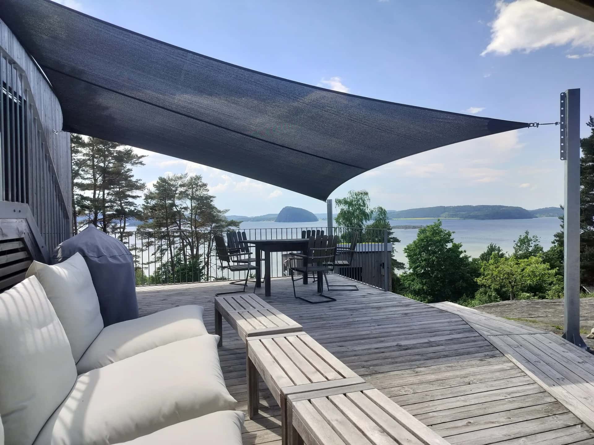Mörkt grått solsegel spännt över träaltan med lounge- och matmöbler vid havsutsikt